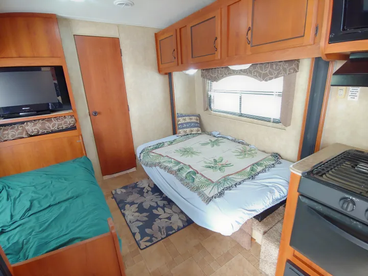 Inside an RV rental: a cozy bedroom with bed, TV, overhead cabinets, and a partial view of the kitchen stove.. Coachmen Catalina 2011