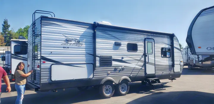 A large, striped gray and white travel trailer RV, possibly for RV rental, with a ladder and awning. A girl stands nearby in an outdoor lot.. Jayco Jay Flight 2021