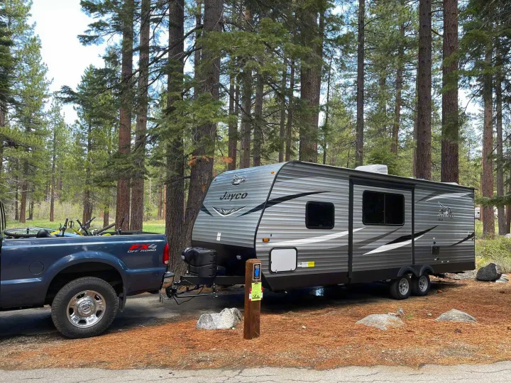 A gray RV rental travel trailer is parked with a blue pickup truck and bicycles at a serene forest campsite.. Jayco Jay Flight 2021