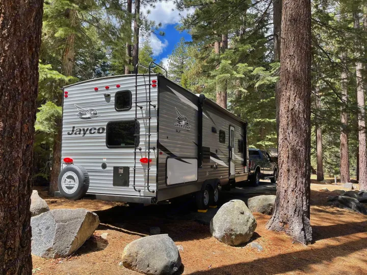 An RV rental, a grey Jayco travel trailer, is parked among tall pine trees and large rocks at a sunny campsite.. Jayco Jay Flight 2021