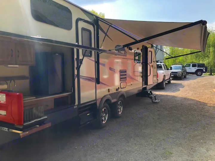 An RV rental with its outdoor kitchen pulled out and awning extended, parked at a sunny gravel campsite with vehicles.. Coachmen 310 BHDS 2015