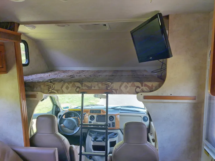 View of an RV rental's interior, highlighting the overhead bunk with ladder, driver's cockpit, and mounted TV.. Winnebago Access 2011