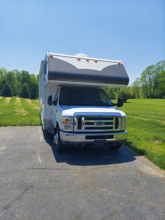Front view of a white Class C RV rental parked outdoors on an asphalt driveway. Bright sunny day with green landscaping.. Winnebago Access 2011