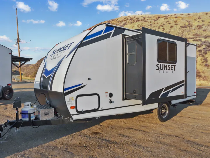 This white travel trailer RV rental, featuring a slide-out, is parked on a dirt lot with hills.. Crossroads Sunset Trail Super Lite 2021