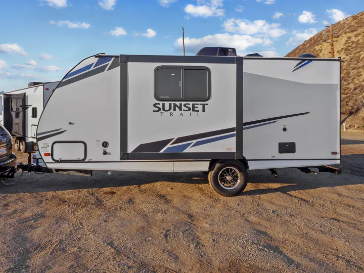 White Sunset Trail travel trailer RV rental parked on a gravel lot with a hill in the background.. Crossroads Sunset Trail Super Lite 2021