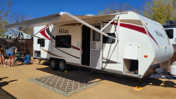 A white travel trailer RV rental with an extended awning, outdoor rug, and people nearby, set up for camping.. Eclipse Recreational Vehicles Milan 2013