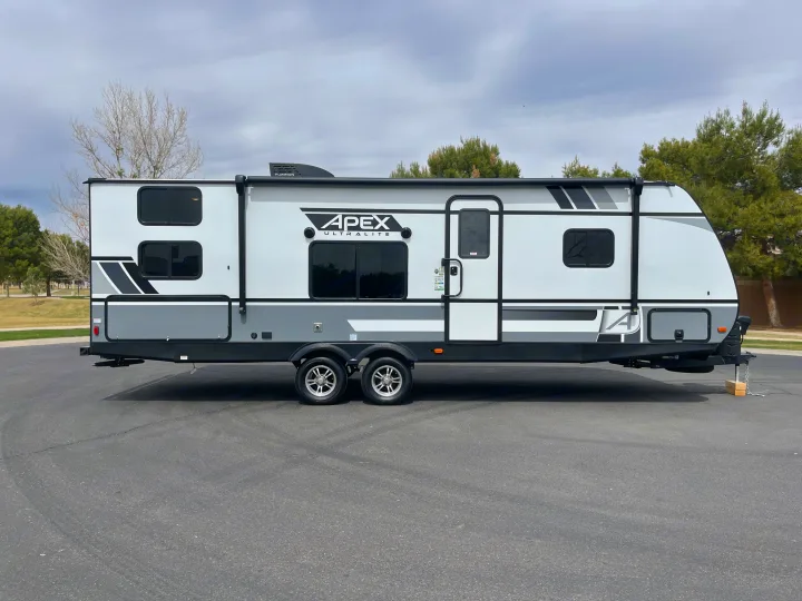 Spacious white and gray travel trailer RV rental parked on asphalt under cloudy skies.. Coachmen Apex 2021