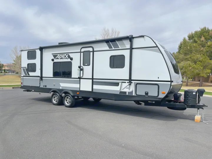 Modern Apex travel trailer for rv rental. Two-axle, white and gray with awning, parked on asphalt.. Coachmen Apex 2021