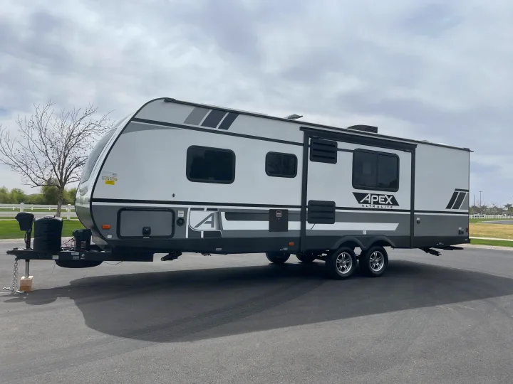 A modern grey and white Apex UltraLite travel trailer, ready for an rv rental, is parked outdoors on asphalt under an overcast sky.. Coachmen Apex 2021