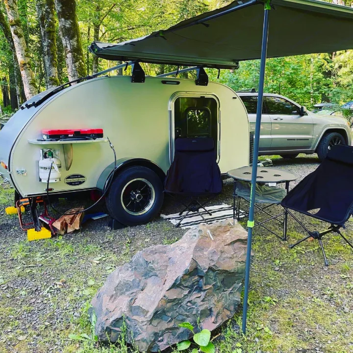 An RV rental teardrop trailer camp setup at a wooded campsite, complete with an awning, table, chairs, and cooking gear.. Aero Teardrops Teardrop Camper 2022