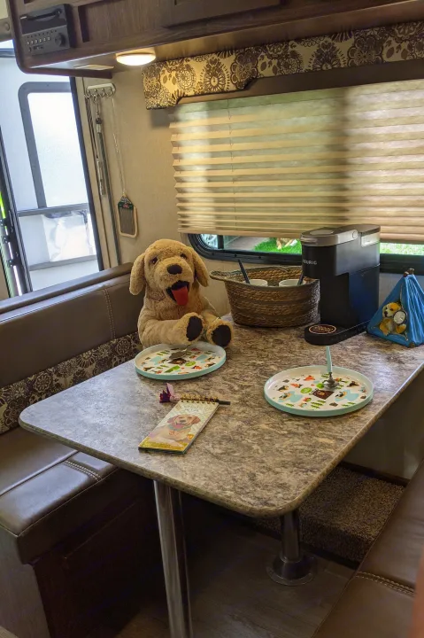 A plush dog sits at an RV rental dinette table with plates & coffee, ready for a cozy journey in this inviting home on wheels.. Other Other 2017