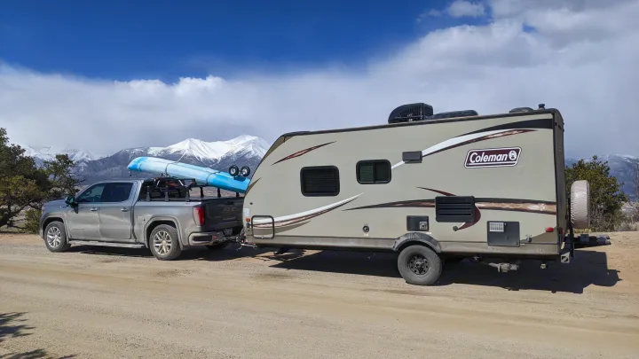 A GMC pickup truck, carrying two blue kayaks, tows a Coleman RV rental on a dirt road with snowy mountains in the background.. Other Other 2017