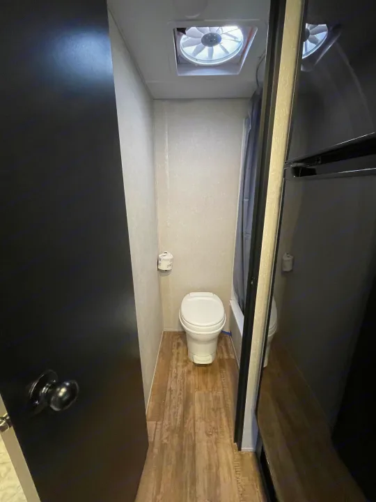 Compact RV rental bathroom features a white toilet, toilet paper on the wall, wood-look floor, and a ceiling vent fan.. Forest River Cherokee Wolf Pup 2021
