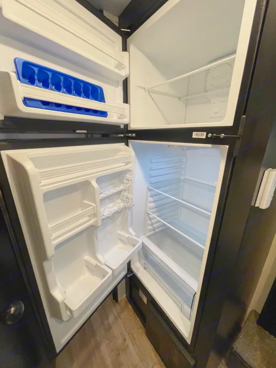 Inside an empty, clean RV rental refrigerator: upper freezer with blue ice trays, lower fridge with adjustable shelves.. Forest River Cherokee Wolf Pup 2021