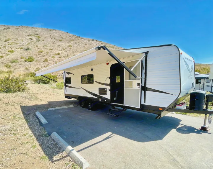 An RV rental, white with an extended awning, is parked on asphalt in a dry, hilly landscape under a clear sky.. Dune Sport Toy Hauler 2017