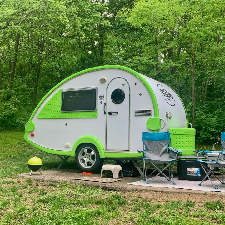 Vibrant green & white teardrop **RV rental** set for camping in a lush forest, with chairs & cooler.. T@B T16 2008
