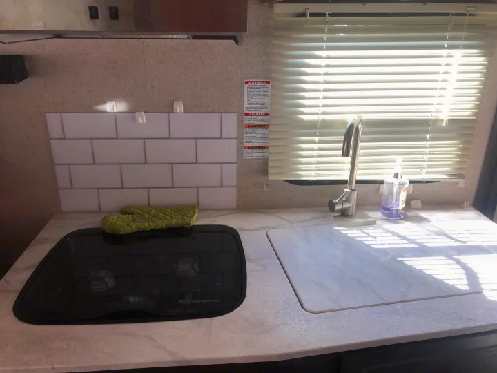 RV rental kitchen: compact counter with stove, sink, and tile backsplash. Propane warnings visible near window.. Dutchmen Kodiak 2019