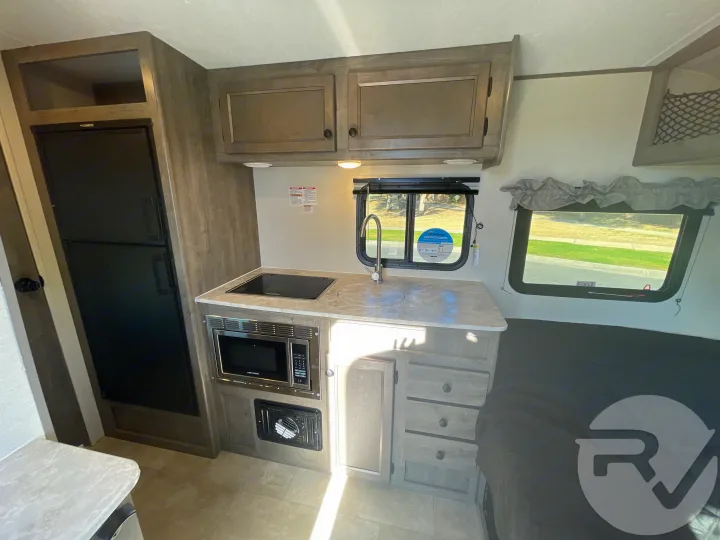 Explore this RV rental's interior, showcasing a compact kitchen with stove, microwave, sink, fridge, and sleeping area.. Coachmen Apex 2021