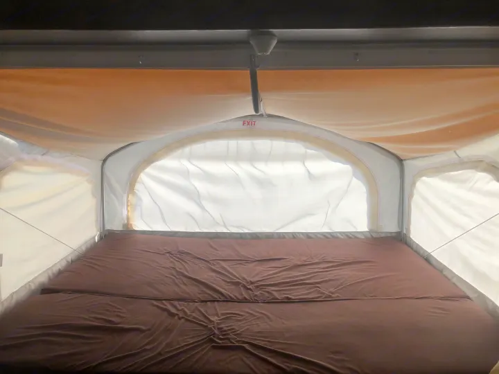 Inside an RV rental: Brown mattress fills this pop-up camper's cozy sleeping area, with fabric walls & exit sign.. Dutchmen Kodiak 2019