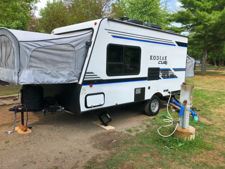 White Kodiak Cub hybrid RV rental set up at a leafy campsite, connected to utility hookups for a comfortable stay.. Dutchmen Kodiak 2019