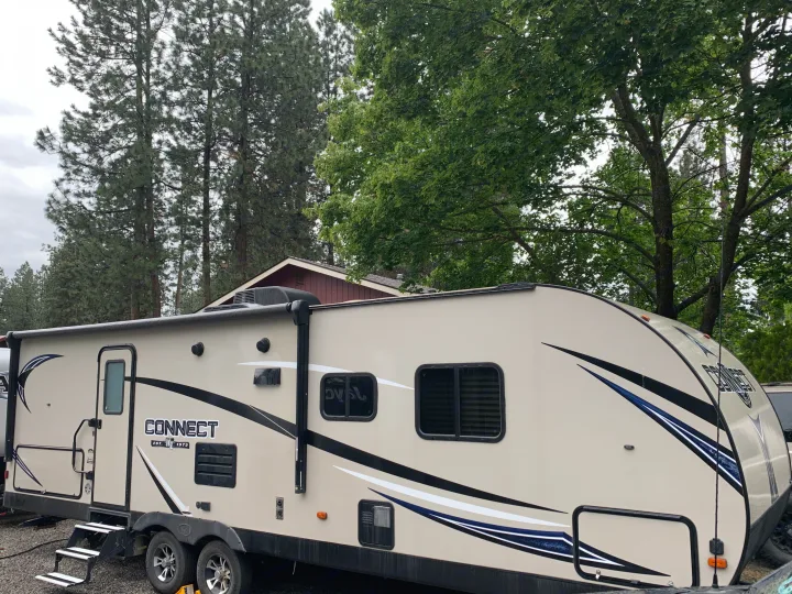 Connect RV rental travel trailer parked outdoors on gravel. Beige body with black, white, and blue stripes. Surrounded by tall trees.. K-Z Manufacturing Connect 2018