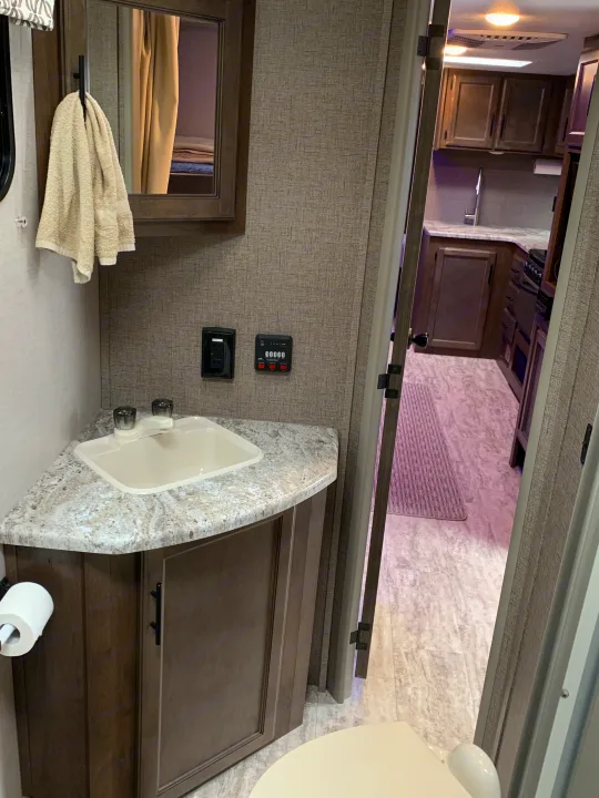 RV rental bathroom featuring a corner sink, mirror, wall controls, toilet, and a glimpse of the kitchen.. K-Z Manufacturing Connect 2018