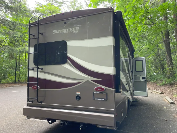 A modern Sunseeker **RV rental** from the rear, showing its ladder & open side door, parked in a leafy forest setting.. Forest River Sunseeker 2019
