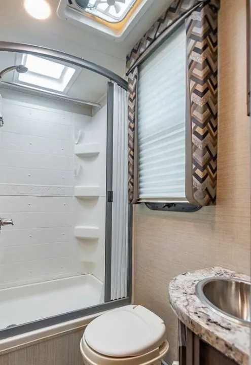 Compact RV rental bathroom with a white shower, toilet, and sink, complete with patterned countertop and window with blinds.. Jayco Redhawk 2018
