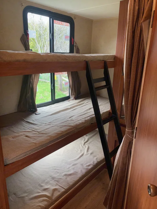 Interior view of an RV rental: three cozy bunk beds with ladder and emergency exit window.. Forest River Surveyor Sport 2014