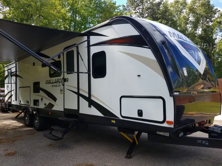 White Mallard travel trailer rv rental with an extended black awning, parked on asphalt with green trees visible.. Heartland M26 2019