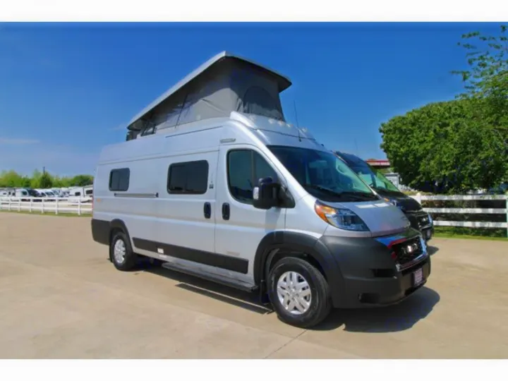Silver pop-top camper van, an RV rental, parked on a paved lot under a clear blue sky, showing its passenger side.. Winnebago Solis 59PX 2022