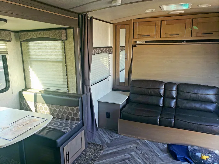 RV rental interior features a dinette, a black leather sofa, and a wall-mounted bed above it. Wood-look floors and ample cabinets.. Cruiser Rv Corp Shadow Cruiser 2020