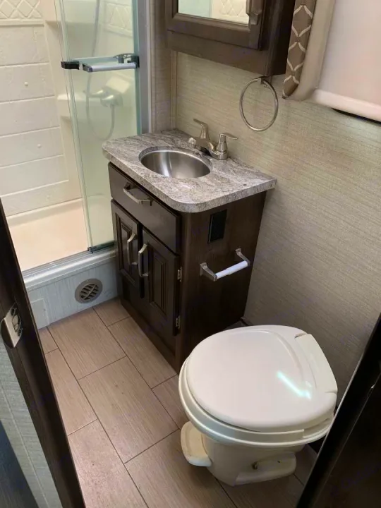 This RV rental bathroom features a glass-enclosed shower, dark wood vanity with a stainless sink, and a white toilet on light wood-look tile.. Jayco Greyhawk 2020