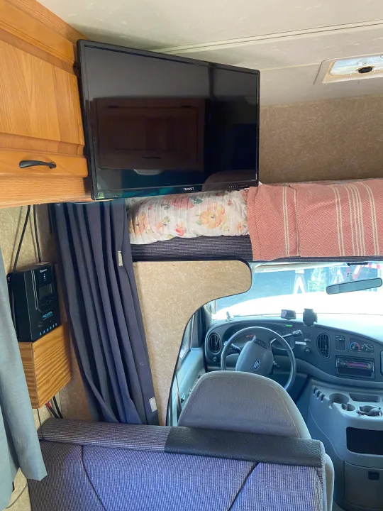 Inside an RV rental: TV mounted above a sleeping bunk, with the Ford driver's cab visible. A solar charge controller is on the left.. Thor Motor Coach Four Winds Majestic 2009