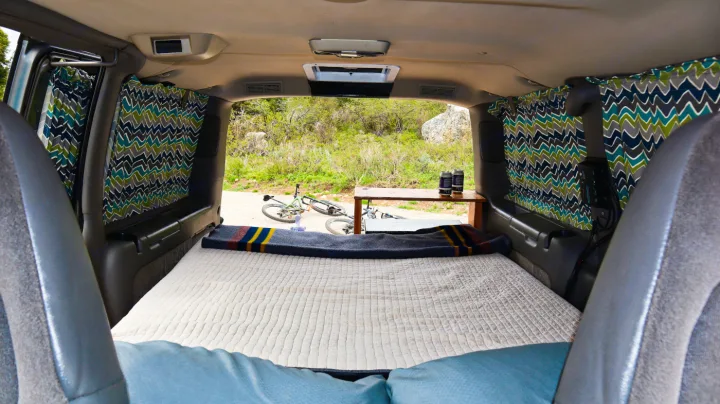 Interior of an RV rental camper van: a bed with striped blanket, patterned curtains, and bikes outside.. Chevrolet Other 2003