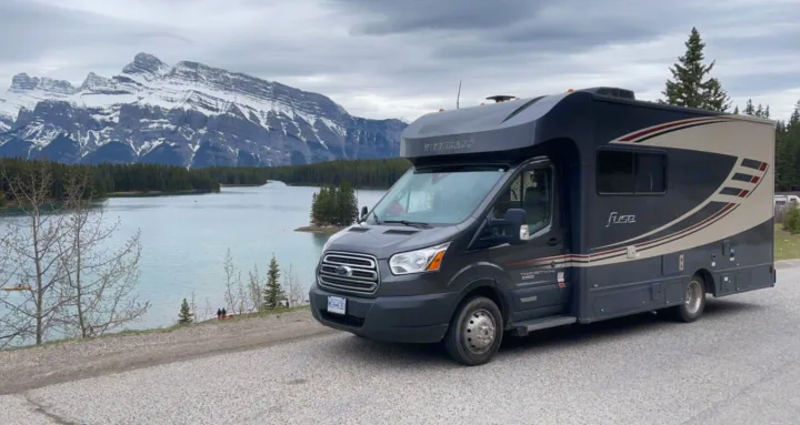 A dark grey Winnebago Fuse RV rental by a serene mountain lake, surrounded by snow-capped peaks and green forests.. Winnebago Winnebago 2018