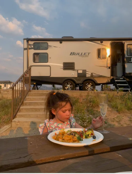 A young girl enjoys dinner at a picnic table during an RV rental trip, with a Bullet Ultra Lite travel trailer behind her.. Work and Play Work and Play Trailer 2020
