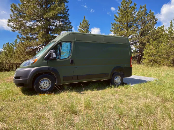 Explore the outdoors with this green RV rental van, nestled in a sunny, grassy clearing among pine trees.. Ram Promaster 2020