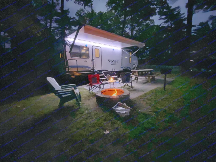 Cozy night at an RV rental campsite. Illuminated RV, warm campfire, chairs, and picnic table under a dark sky.. Forest River Vibe 2021