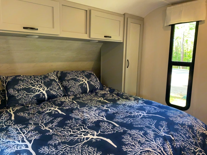 Cozy RV rental bedroom featuring tree-patterned bedding, overhead cabinets, and a window with a nature view.. Forest River Vibe 2021
