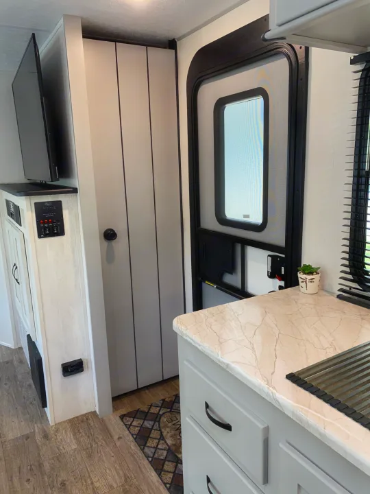 Inside a modern RV rental: a light marble-look kitchen counter, main entry door, light grey cabinets, TV, and wood-look floor are visible.. Forest River Vibe 2021