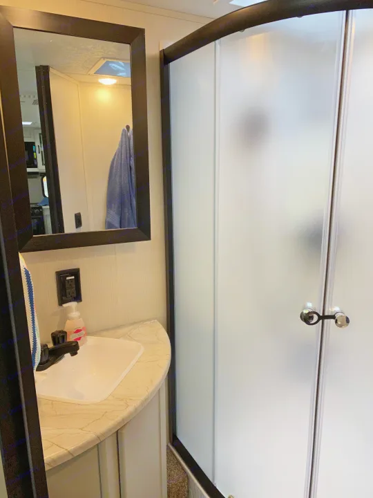 An RV rental bathroom featuring a compact vanity with sink, mirror, and a frosted glass shower with black trim. Efficient for travel.. Forest River Vibe 2021