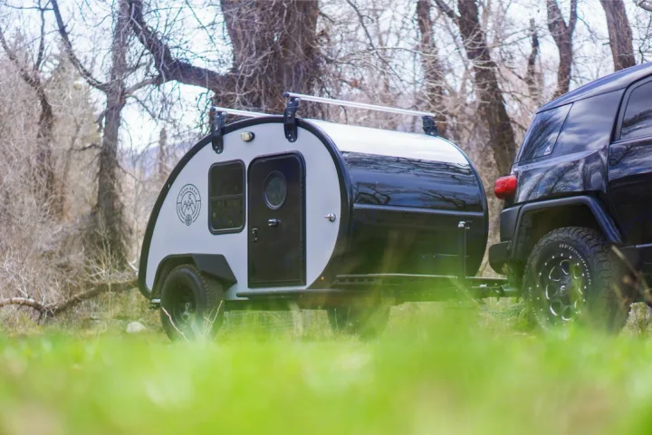 Adventure awaits! This black SUV tows a modern teardrop RV rental, parked in a lush, wooded outdoor setting.. Bean Trailer Mean Bean 2021