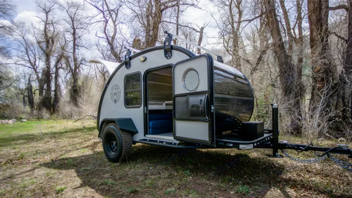 Light grey teardrop RV rental parked in a grassy, wooded area with bare trees and open doors, ready for camping.. Bean Trailer Mean Bean 2021