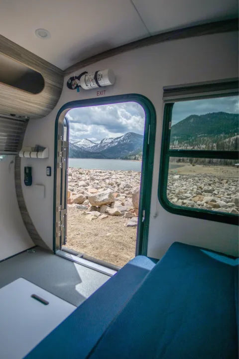 RV rental interior with open door to a stunning lake, snow-capped mountains, & rocky shore. Fire extinguisher above.. Bean Trailer Mean Bean 2021