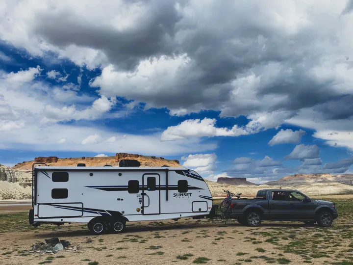 White RV rental travel trailer hitched to a pickup truck in a vast, dry landscape with mesas under a cloudy sky. Ideal for road trips.. Crossroads RV Sunset Trail Super Lite 2021