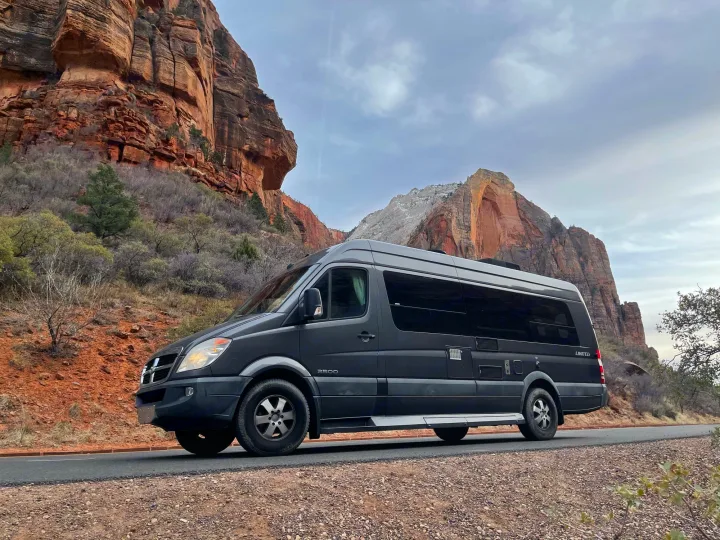 A sleek black RV rental van on a scenic road, flanked by majestic red rock formations under an overcast sky.. Winnebago Era 2010