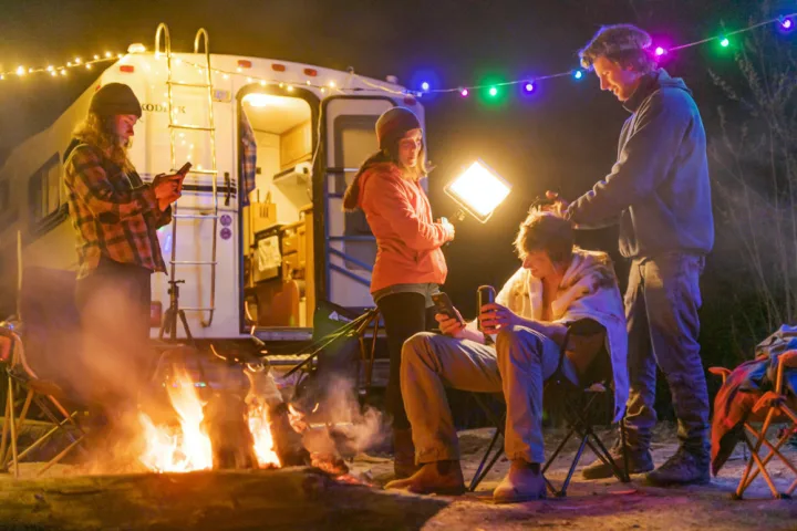 Friends gather around a crackling campfire by their RV rental at night, lit by string lights. Ideal for outdoor adventures.. Ford Other 1996