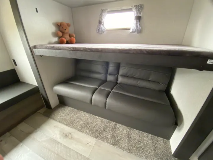 Inside an RV rental: a cozy top bunk with a teddy bear & window, and a gray sofa below for seating.. Sportsmen LE 2021