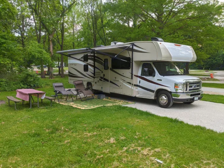 This RV rental is perfectly set up at a wooded campsite, featuring an extended awning, chairs, and a picnic table.. Coachmen Leprechaun 2018